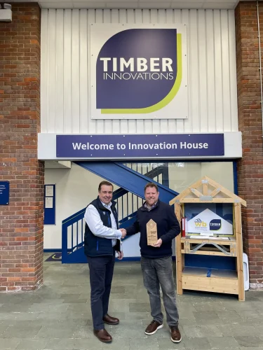 2 men shaking hands in front of a Timber Innovations sign, the man on the right is holding a wooden trophy