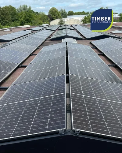 Multiple rows of rooftop solar panels on a commercial roof, trees and buildings in the background, Timber Innovations logo.