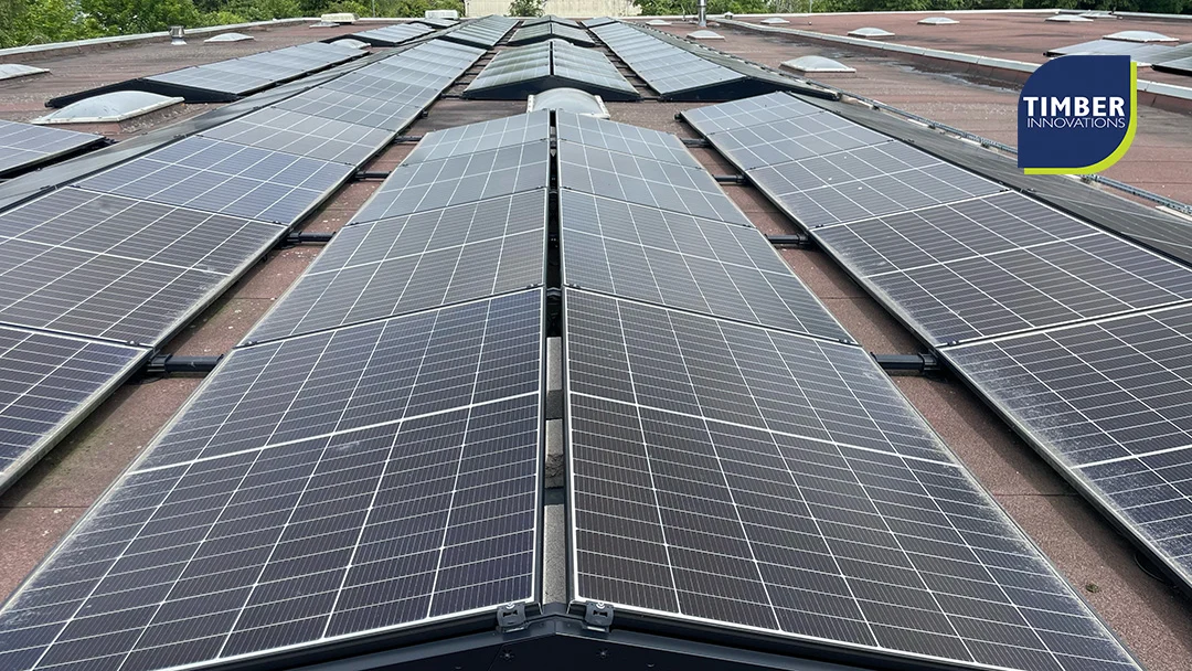 22:06 TI PV 2 Multiple rows of rooftop solar panels on a commercial roof, trees and buildings in the background, Timber Innovations logo.