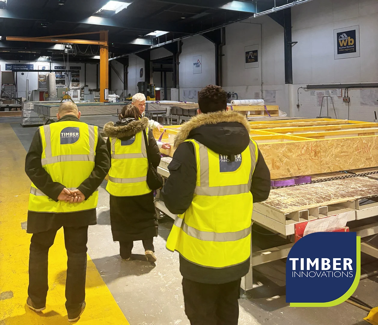 Self-Build-Workshop-Updated- Three people in Timber Innovations high-visibility vests touring a timber factory, inspecting wooden frame panels on worktables.